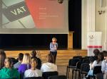 Edukatorka akademii podatkowej podlaskiej KAS prowadzi lekcję dla uczniów I LO w łomży w auli, w tle rzutnik z informacjami o podatkach