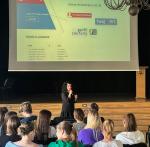 Edukatorka akademii podatkowej podlaskiej KAS prowadzi lekcję dla uczniów I LO w łomży w auli, w tle rzutnik z informacjami o sprawach, które można załatwić w e-US